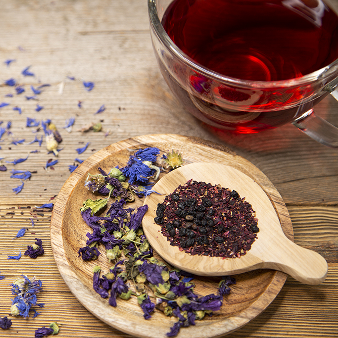 There is a tea in a glass cup on a wooden table next to a bowl with colorful dried flowers and a tea blend
